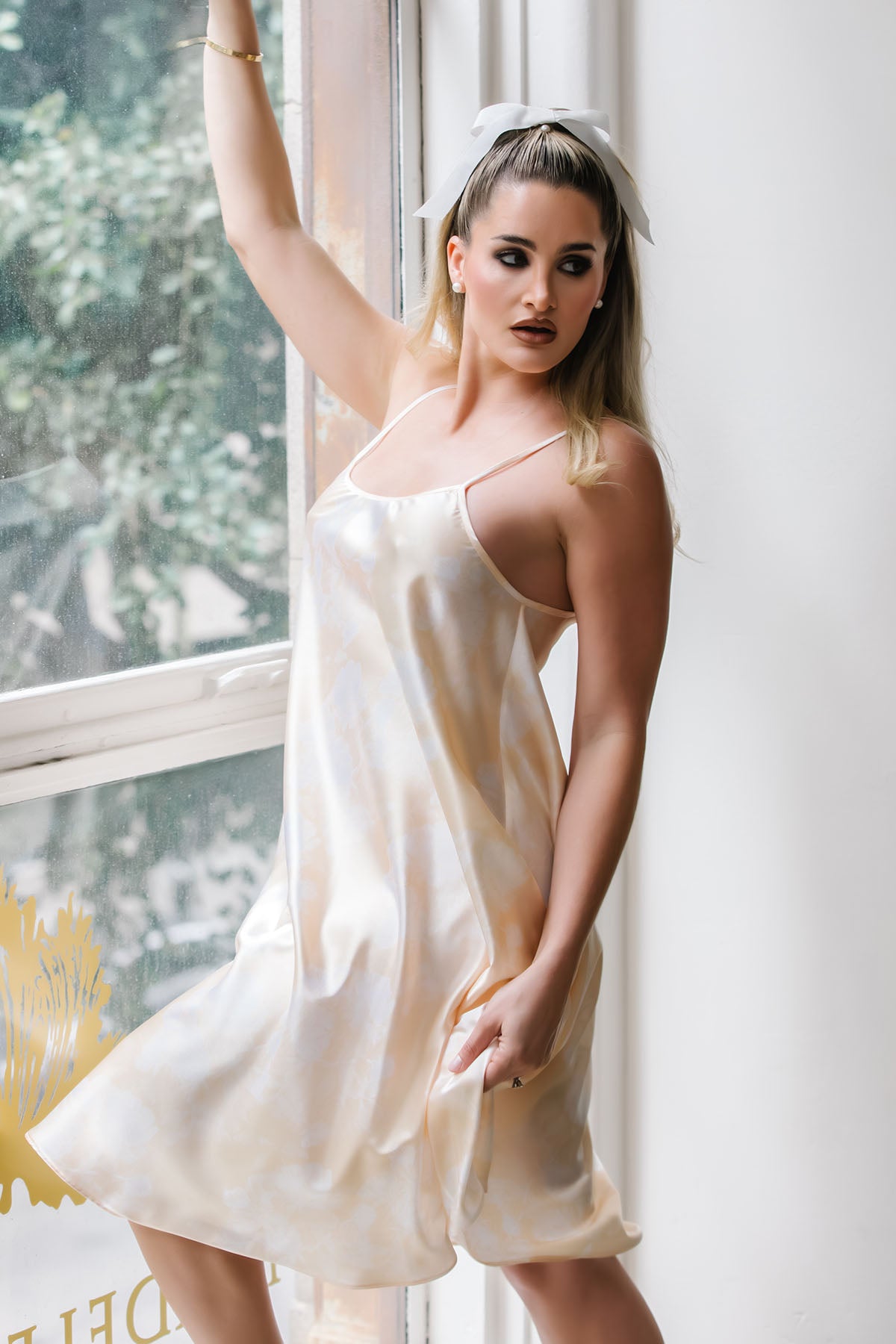 Woman in a white satin slip dress standing by a window