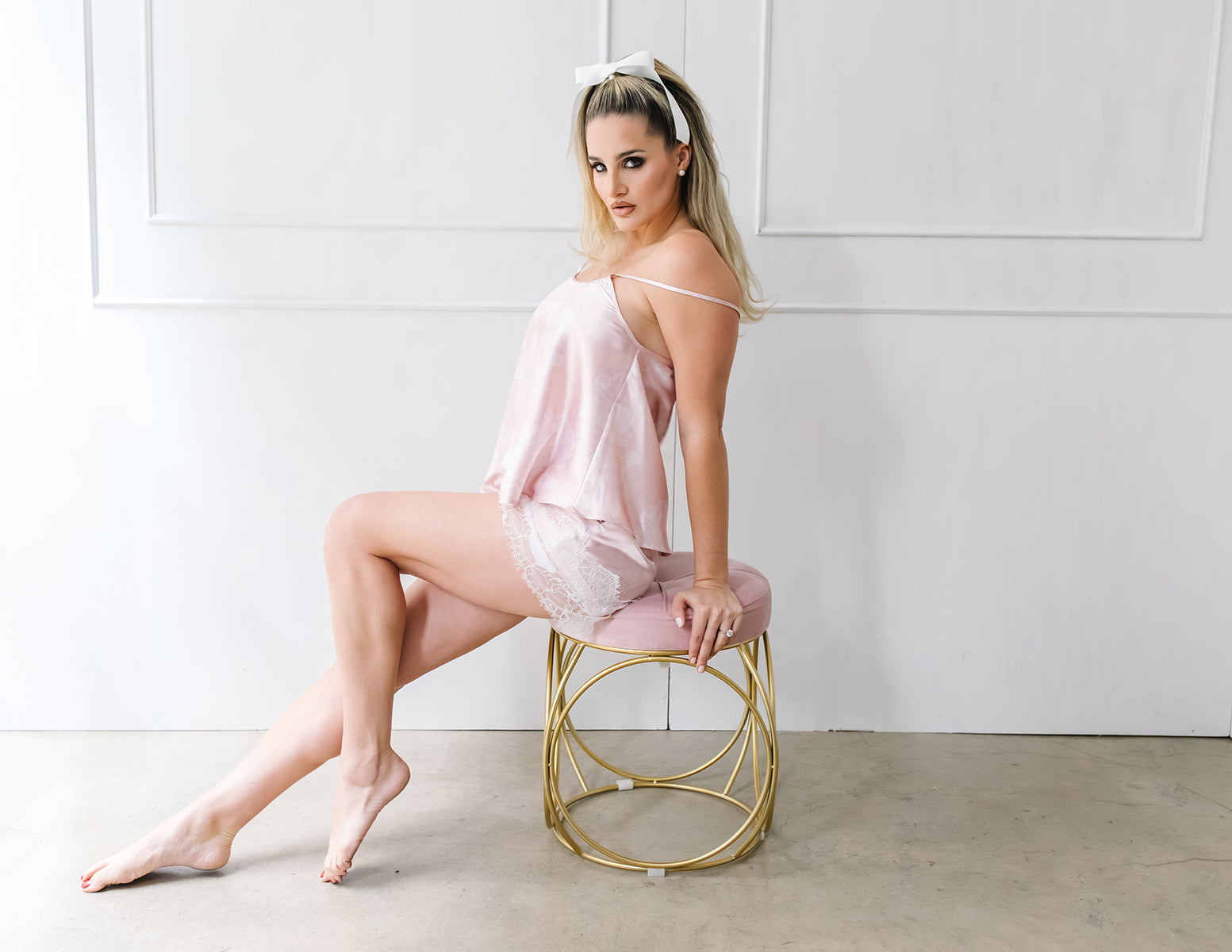 Woman in a pink satin slip sitting on a gold stool against a white background