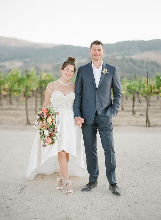 Couple in wedding attire standing in a vineyard with mountains in the background