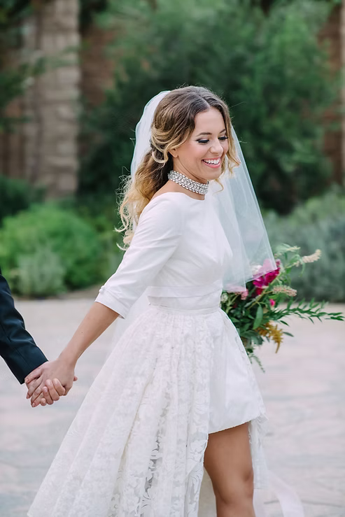 Bride in a white wedding dress holding hands with a groom, surrounded by greenery.