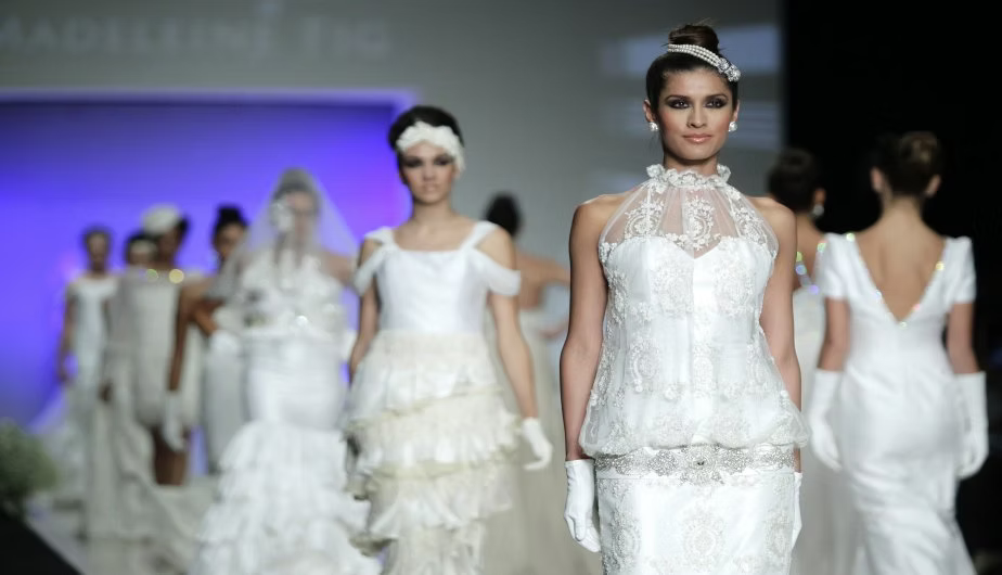 Model wearing a white lace dress on a fashion runway with other models in the background.
